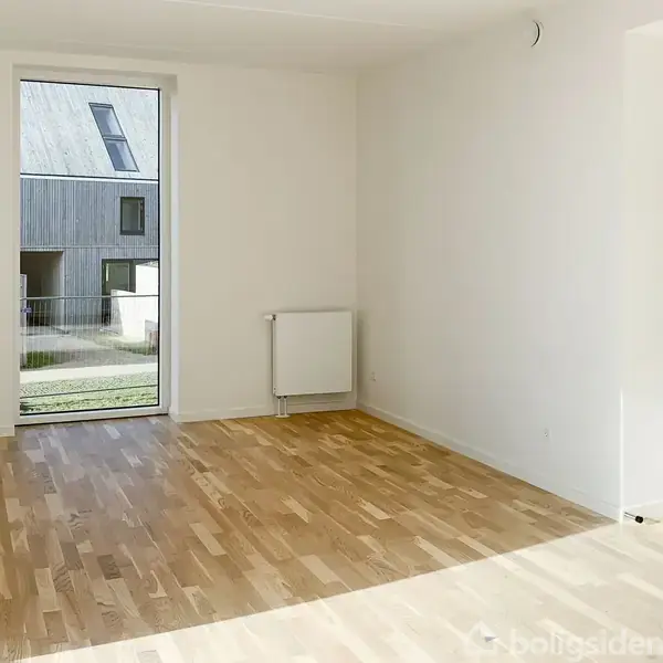 Et lyst rum med trægulv og en stor vindue, der giver udsigt til en bygning udendørs. Væggene er hvide, og der er en radiator under vinduet.