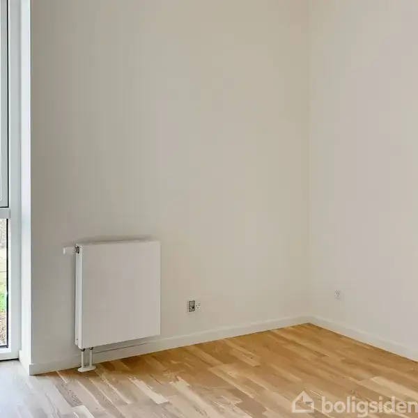 Et tomt rum har hvide vægge og lyst trægulv. En radiator er monteret på væggen. Store vinduer til venstre viser et hus og grønne områder udenfor.