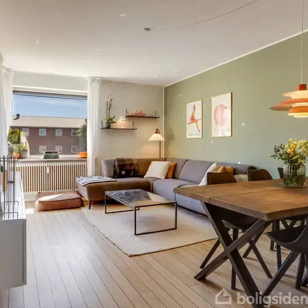 Stue med lys sofa og sofabord placeret på tæppe. Bord med stole og blomster i vase ved siden. Grøn væg med kunst. Store vinduer giver naturligt lys.