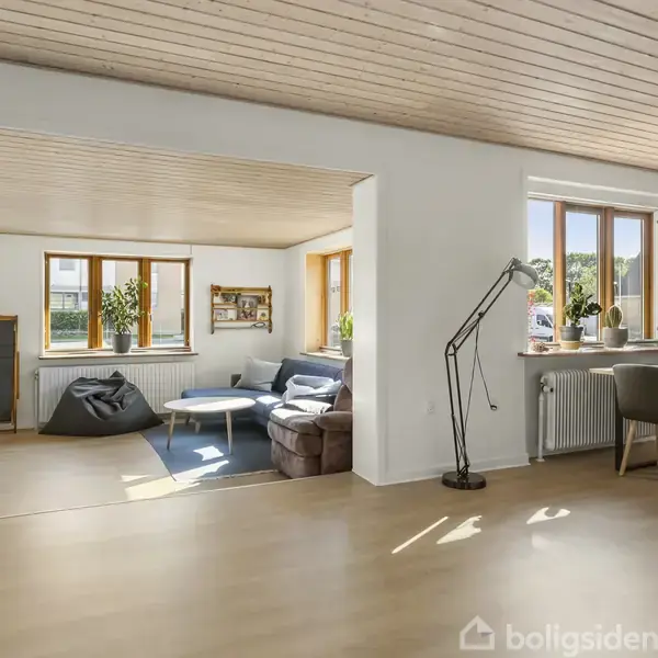 En stue med en grå sofa og lænestol ved siden af et vindue. I baggrunden ses et spisebord med stole. Trægulve og lyse trælofter skaber en lys atmosfære.