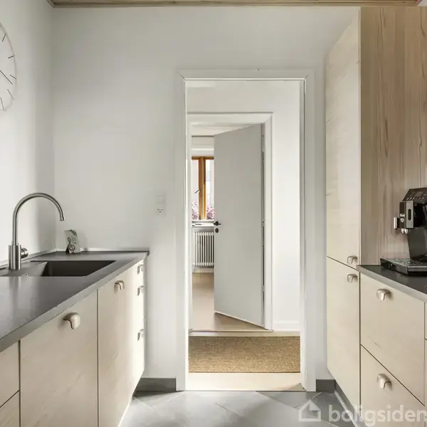 En køkken med lyse træskabe og en mørk bordplade. En vask og en kaffemaskine er synlige. Et ur hænger på væggen. Døren fører til et lyst rum.