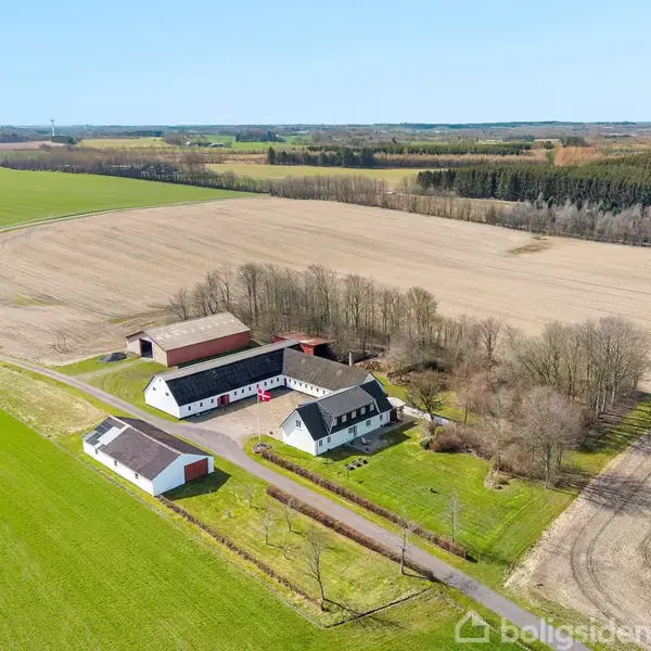 Gårdbygning ligger stille omgivet af åbne marker og skov. Flere bygninger ses langs en vej med grønne arealer og marker omkring.