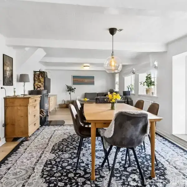 Et spisebord med stole står i et lyst rum med hvidt loft og vægge. Store vinduer til højre lukker naturligt lys ind, mens en dekorativ tæppe pryder gulvet.