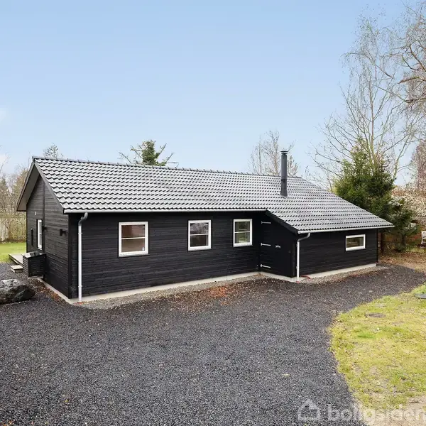 Et sort, enkelt hus står stille på en grusvej omgivet af træer og buske. En lille græsplæne ses ved siden af huset. En skilt med ordet "Til salg" står foran.