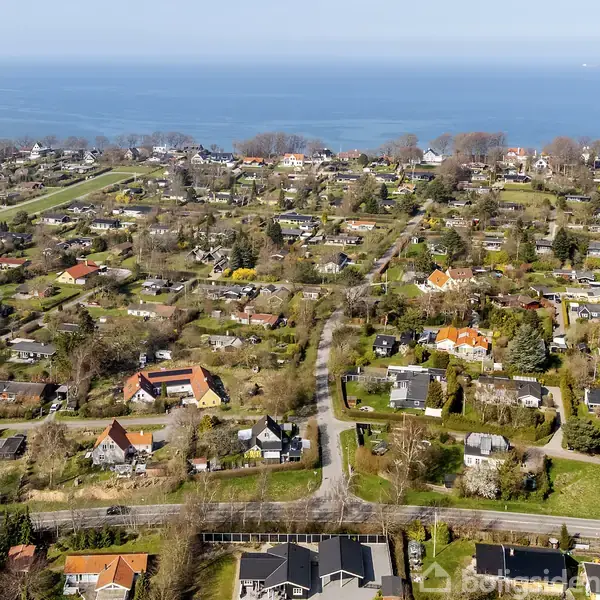 Boligområde set ovenfra med huse spredt ud i et grønt landskab. Baggrunden viser havet, der strækker sig til horisonten. Træer og veje gennemskærer området, der er stille og fredfyldt.