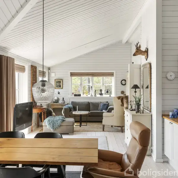En lys stue med trævægge har en træspisebord og sorte stole. Sofaer står ved vinduet. Hængelamper over bordet. Et blødt tæppe og lammehoved pynter rummet.