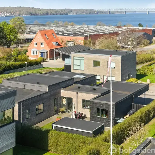 Flere moderne, kubiske huse med fladt tag og store vinduer står side om side. Et rødt gavlhus ses i baggrunden. Landskabet afsluttes med en bro og en vandudsigt.