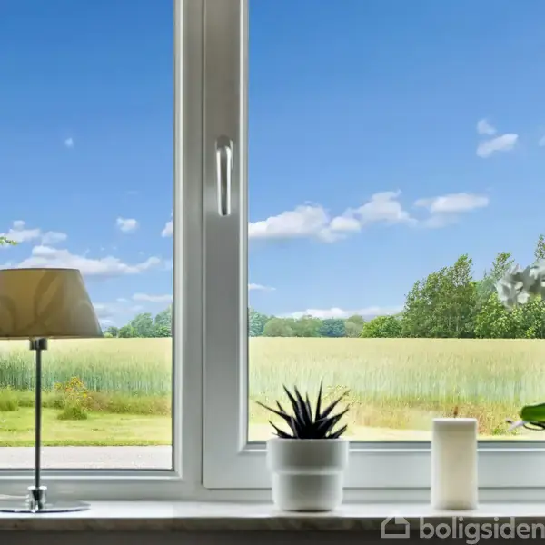 Vinduet har udkig til en mark med træer og blå himmel. En lampe, en sukkulent, et stearinlys og en orkidé står på vindueskarmen, hvor dagslys strømmer ind.