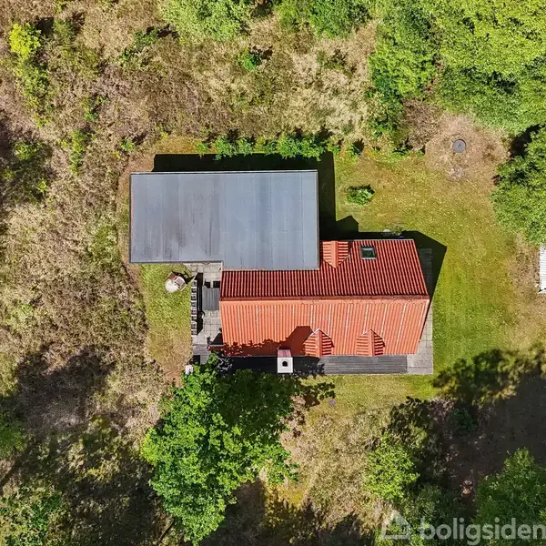 Hus med rødt tag set oppefra, omgivet af græsplæne og træer, placeret i et skovområde. Der er en terrasse ved huset, og området er skyggefuldt og grønt.