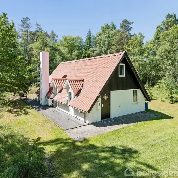 Et hus med rød tag og hvid facade står på en græsplæne omgivet af skov. Huset har små vinduer, en høj skorsten og en terrasse ved siden af.