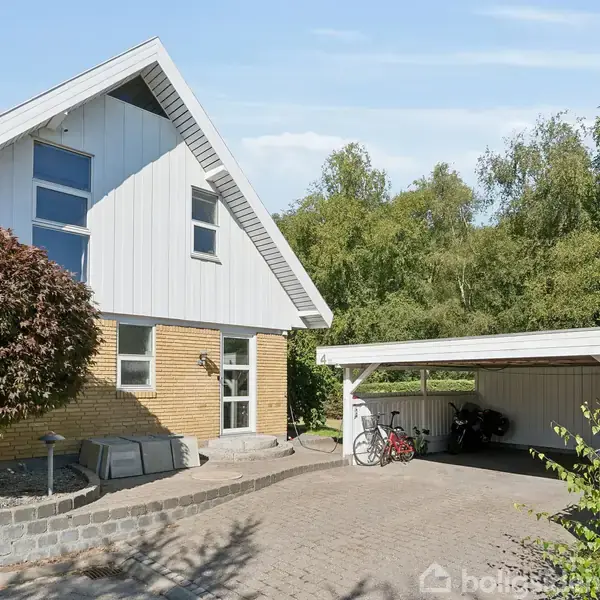 Et moderne, to-etagers hus med murstensfacade og store vinduer står ved siden af en carport med cykler. Omgivet af træer og en brolagt indkørsel i et fredeligt boligområde.