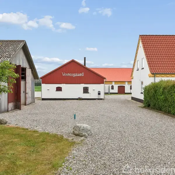 En gårdsplads med grus i midten, omgivet af flere bygninger med røde tage. Bygning i midten har teksten "Vestergård". Grønne buske og et træ er synlige til venstre.