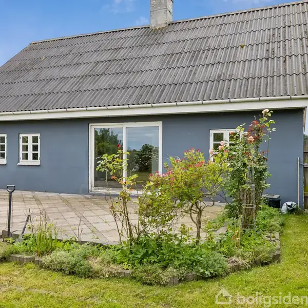 Hus med gavltag står stille i en grøn have med flisebelagt terrasse. Omgivet af buske og blomster, kompleteret af blå himmel baggrunden.