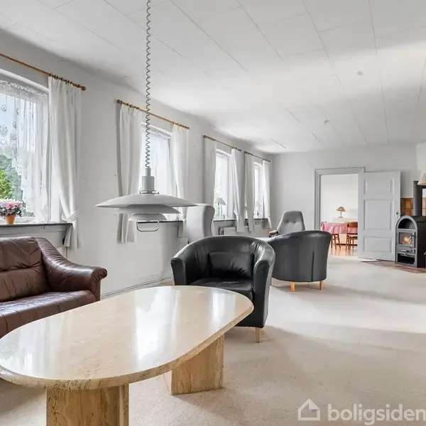 Brune lædermøbler står i en lys stue med store vinduer og hvide gardiner. Et sofabord er centralt placeret, med en lampe hængende over. En brændeovn ses i baggrunden.