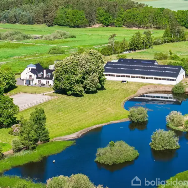 En hvid bygning og et stort skur står ved siden af en dam omgivet af åbne grønne marker og træer. I baggrunden ses tæt skov.