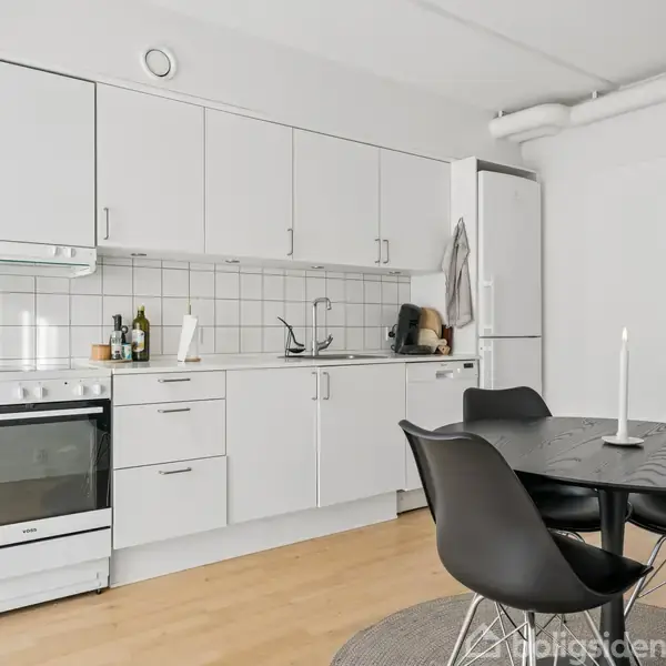 Et moderne køkken har hvide skabe, komfur og opvaskemaskine. Sort bord med sorte stole står på et rundt tæppe. Fliser som backsplash tilføjer stil til det lyse rums udtryk.
