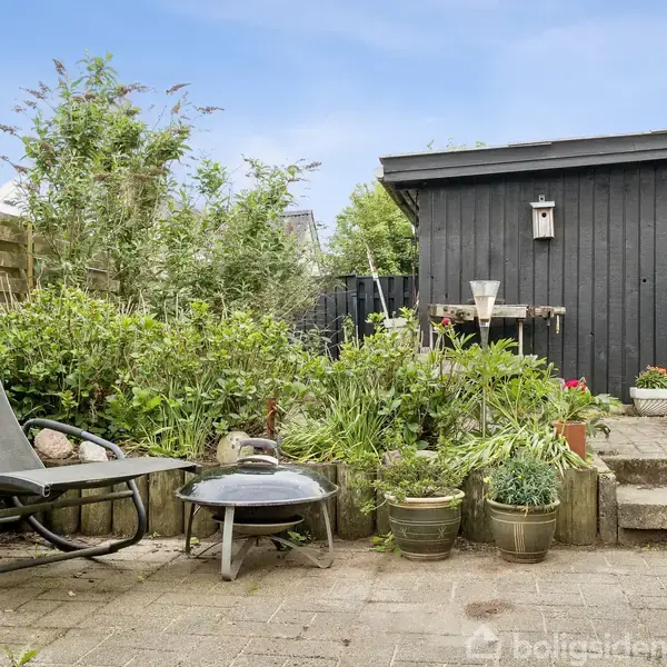 En sort liggestol står på en flisebelagt terrasse ved siden af et lavt bord og en lille have med frodige planter. I baggrunden ses en sort træbygning.