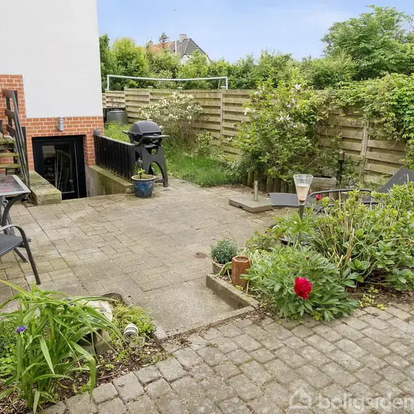 Terrasse med borde og stole, placeret ved siden af en bygning. Området har en grill, omgivet af blomster, planter og en have, med en træhegn bagved.