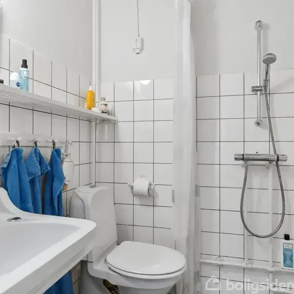Håndvask med spejl og hylde over, placeret til venstre. Toilet i midten ved siden af en håndklædestang med blå håndklæder. Bruser til højre i et flisebelagt badeværelse.