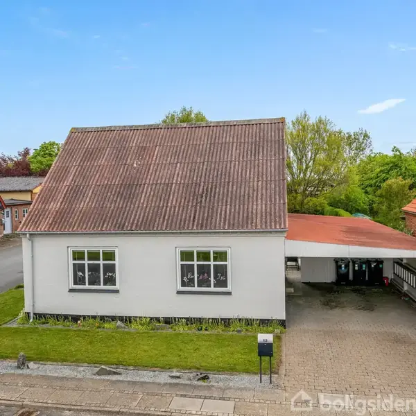Hvidt hus med rødt tag, to vinduer; står langs en villavej. Indkørsel med overdækket carport til højre. Græsplæne foran omgivet af hække og nabohuse.