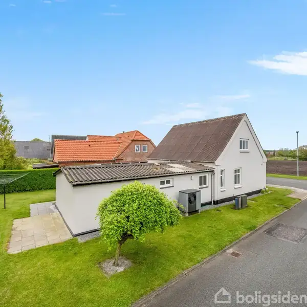 Et hvidt hus med en skrånende tag og et træ ved siden står ved en vej. Græsplæne og marker omringer huset under en blå himmel.