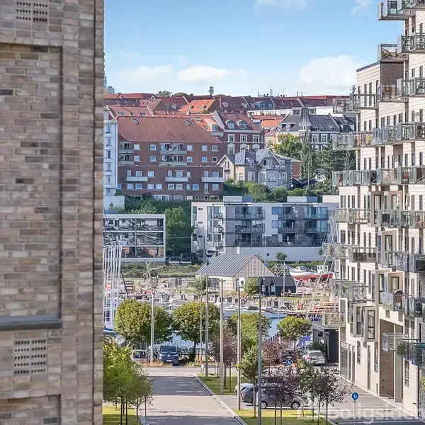 Et område med moderne lejlighedsbygninger omgiver en vej med træer og parkerede biler; i baggrunden ses en havn og bakker med rødtagsbygninger.