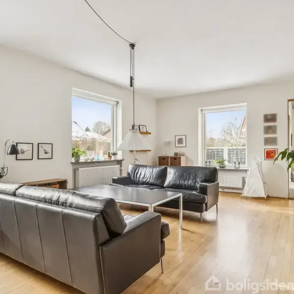 To sorte lædersofaer står omkring et sofabord i en lys stue med trægulv. Store vinduer giver udsigt til træer udenfor. Flere planter og kunstværker dekorere rummet.