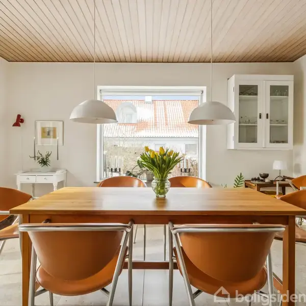 Et spisebord med gule blomster i en vase står centralt. Omgivet af seks brune stole, mens to lamper hænger over bordet. Bagved ses et vindue og en skænk.