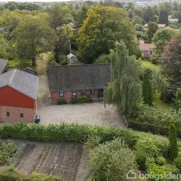 Rødt murstenshus og lade står stille omgivet af grønne træer og buske. Grøntsagsbed ses i forgrunden. Bag ved bygningen anes flere huse og skove i horisonten.