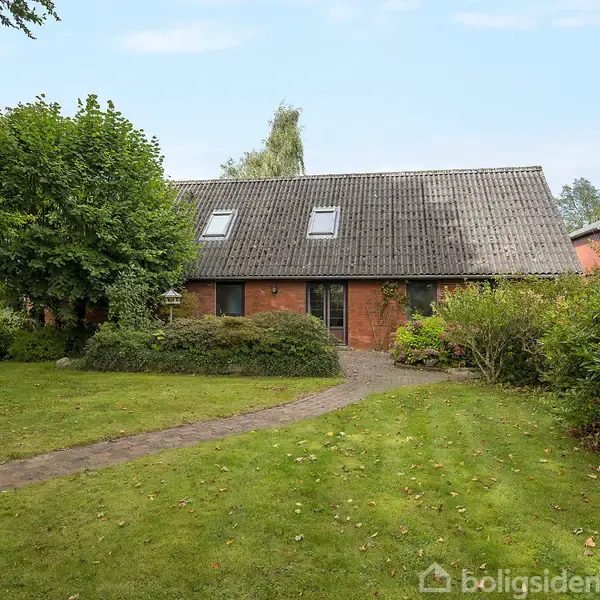 Et rødt murstenshus med sort tag og to ovenlysvinduer står omgivet af grønne buske og træer i en velplejet have med en græssti, under en klar blå himmel.