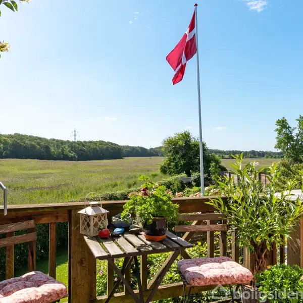 En terrasse med træmøbler og planter står i forgrunden. Et dansk flag vajer i vinden. I baggrunden ses en grøn mark og skov under en skyfri himmel.