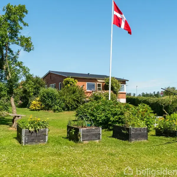 Et dansk flag vajer på en flagstang foran en rød murstensbygning i en grøn have. Flere plantekasser står på græsset, mens et tørrestativ ses i baggrunden.