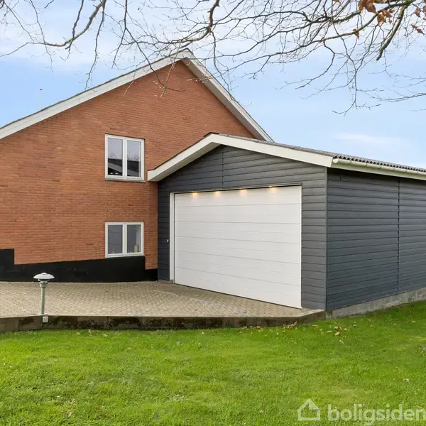 Rødt murstenshus med en moderne, grå garagebygning, står på en velplejet græsplæne. Omgivelserne er delvis træer og åbent landskab under en klar himmel.