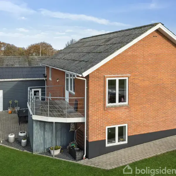 Rødt murstenshus med sort tag i to etager, står på en velplejet græsplæne ved siden af en grå garage. Foran er en terrasse med potteplanter, omgivet af en brostensbelagt gangsti.