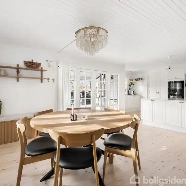 Rundt spisebord med fire stole står i et lyst, åbent køkken-alrum. Der er hylder med dekorationer på væggen, store vinduer og en gulv-til-loft glasdør, samt en elegant lysekrone.