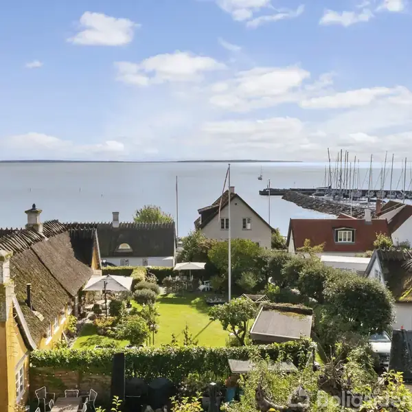 Charmerende landsbyhuse med stråtage står stille i en grøn have, omgivet af frodig vegetation. I baggrunden ses en marina fyldt med både, og der er udsigt over en rolig bugt.