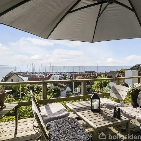 Terrasse med træmøbler, store parasol og indrettet med planter og lanterne. Udsigt over en lille by med havneområde, mange både og havet i baggrunden under en blå himmel.