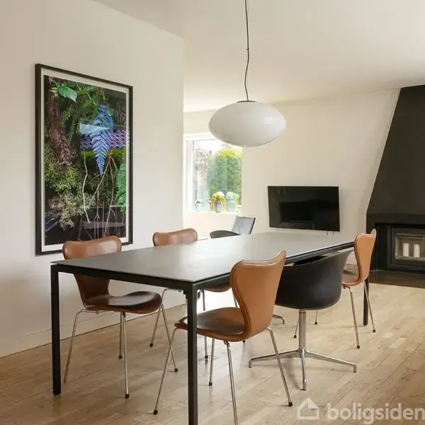 Et moderne spisebord med seks stole står i et lyst rum. En stor lampe hænger over bordet. Væggen er dekoreret med farverigt kunsttryk. En pejs står i hjørnet.