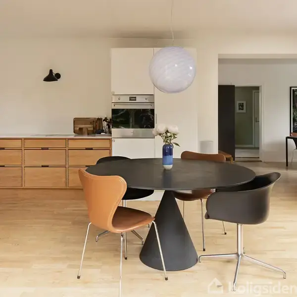 Et moderne køkken med lys trægulv har et rundt sort spisebord omkranset af forskellige farvede stole. Loftlampe hænger over bordet. Køkkenskabe og ovn ses i baggrunden.