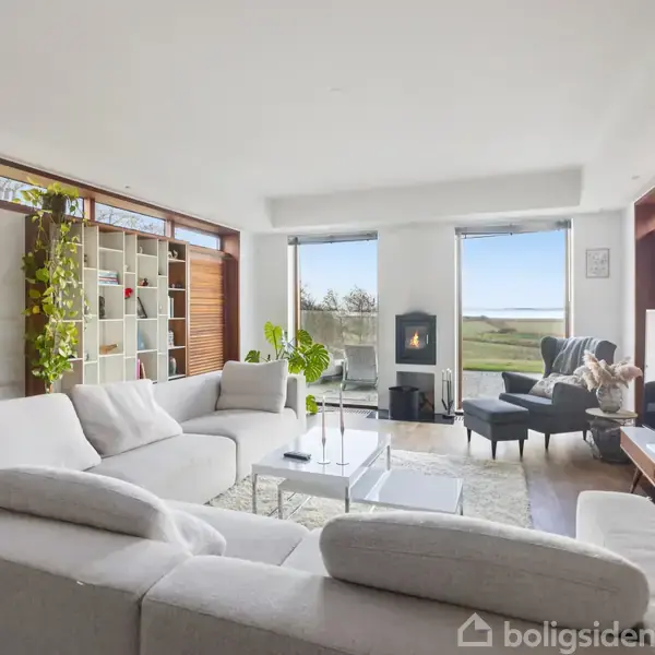 En lys stue med store vinduer har en hvid hjørnesofa, der vender mod et fladskærms-TV. En pejs står ved siden af lænestole, mens reoler med planter dekorerer rummet.