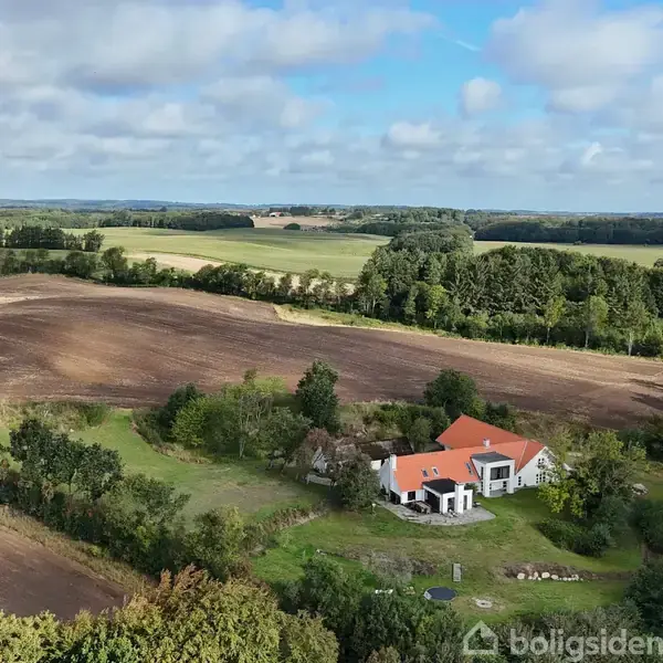 Hus med rødt tag ligger i en grøn have omgivet af skove og marker. En vidstrakt horisont strækker sig under let skyet himmel.