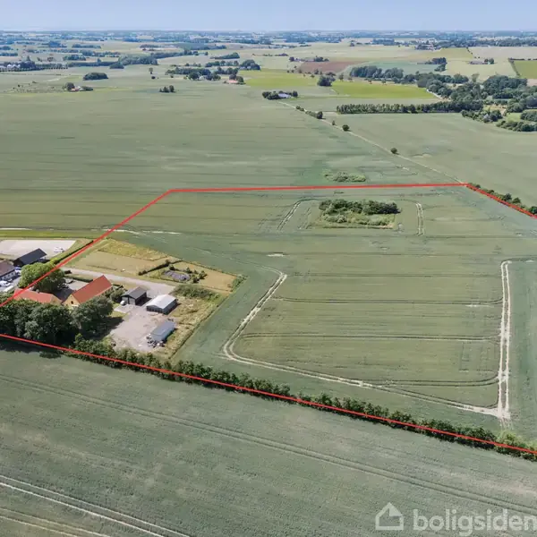 En gård med flere bygninger omgivet af marker, indhegnet med en rød linje. Landskabet er grønt og fladt med spredte træer og vidtrækkende udsigt over marklandskab.