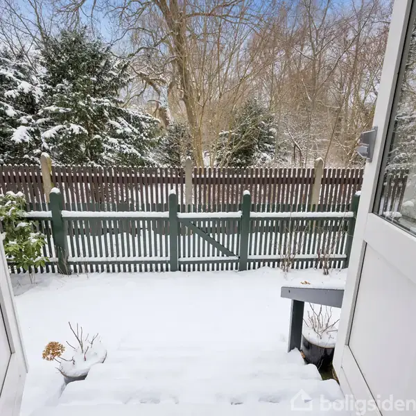 Åben dør med udsigt til en snedækket baghave. Sne dækker trappen, med et stakit og sneklædte træer i baggrunden. Klar blå himmel ses gennem træernes grene.