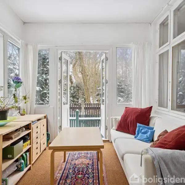 Stue møbleret med sofa, sofabord og skænk. Sofaen har farverige puder. Store vinduer og åbne døre giver udsigt til sneklædt have med træer. Tæppe ligger på gulvet.