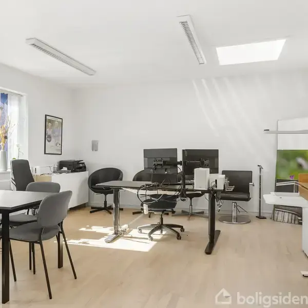 Kontor har skrivebord med computere, kontorstole og skriveudstyr; stole omkring et bord fungerer som mødeområde. Sollys strømmer ind gennem vinduerne, skaber et lyst og luftigt miljø.