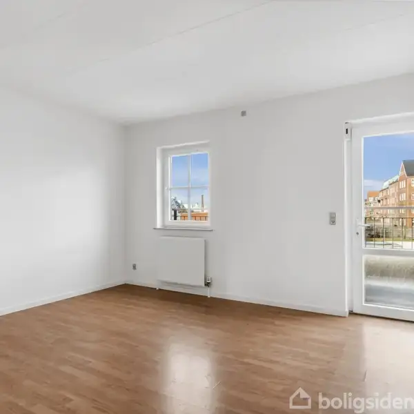 Et tomt rum med trægulv, en radiator under et enkelt vindue, og en glasdør der åbner til en balkon med udsigt til flere bygninger udenfor.