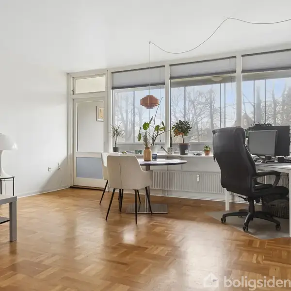 Stue med TV på væggen, klaver og guitar ved siden af sofa. Et sofabord står i midten med frugtskål. Vægdekorationer og en plante skaber en hyggelig atmosfære.