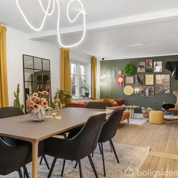 Et moderne spisestueområde med et stort bord omgivet af seks sorte stole, blomster i en vase på bordet. I baggrunden er stuen med kunst på væggen og farverige møbler.