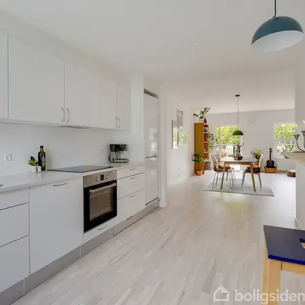 Et lyst køkken med hvide skabe og integreret ovn strækker sig mod en stue. Bord og stole står ved et vindue, med planter og kunst på hylder.