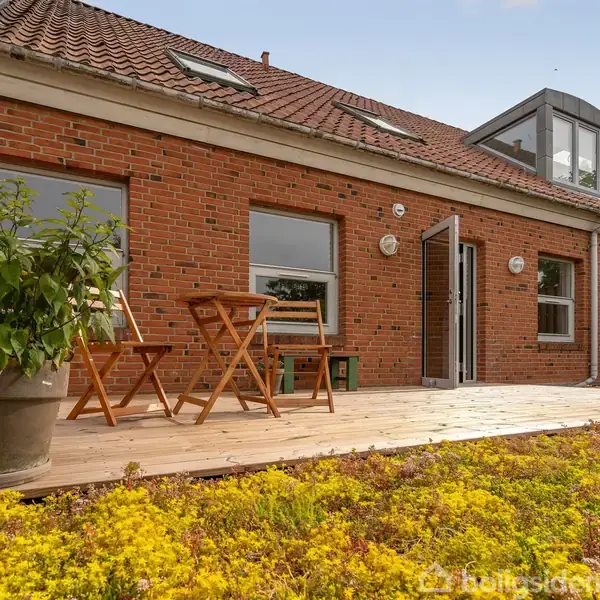 Rødt murstenshus med skråt tag har åbne vinduer og en træterrasse. To træstole og et lille bord står på terrassen omgivet af grønne planter i krukker.